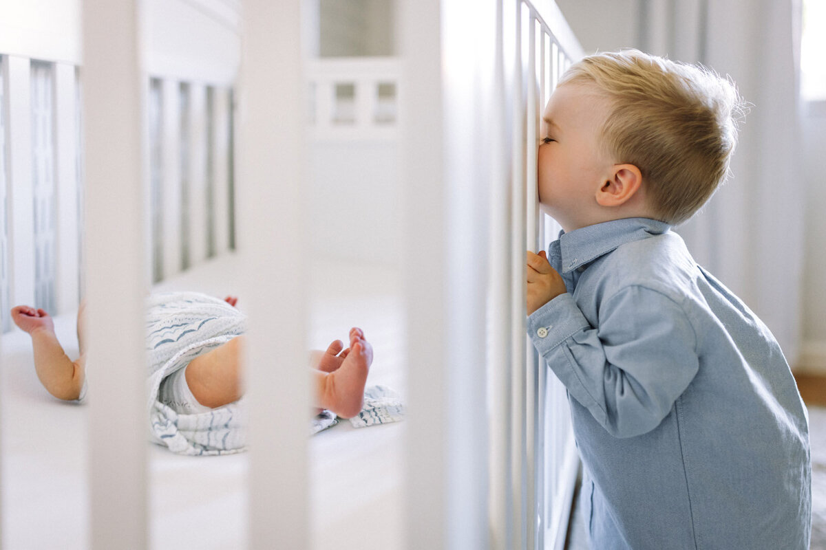 toddler boy in blue button down shirt peeking into white crib to see newborn feet