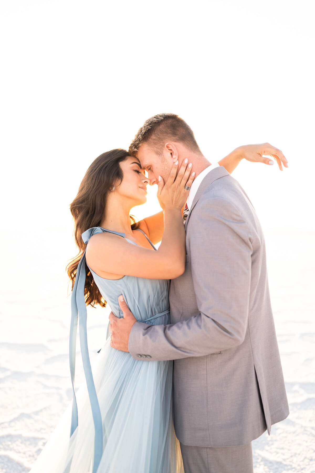 A woman dressed in a soft blue chiffon gown stands caressing her fiancee's face dressed in a grey suit in the Salt Lake Sand Dunes photographed by Bay area photographer, Light Livin Photography.