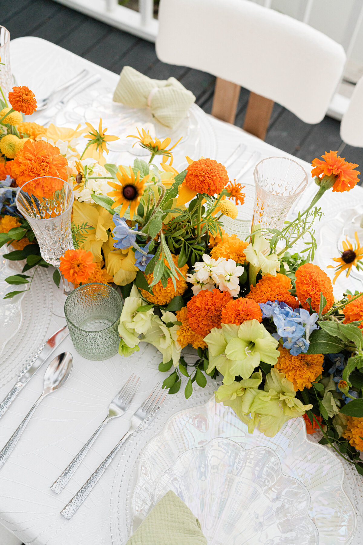 white on white palm leaf tablecloth with hammered silver flatware and an iridescent seashell plate adorned with a white, orange, blue and green floral table runner