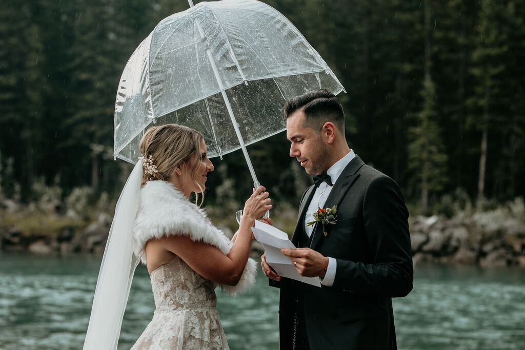 Bride and groom holding clear umbrella during their rainy mountain elopement captured by Lewis and Company, timeless and artful wedding photographer and videographer in Calgary, Alberta. Featured on the Bronte Bride Vendor Guide.
