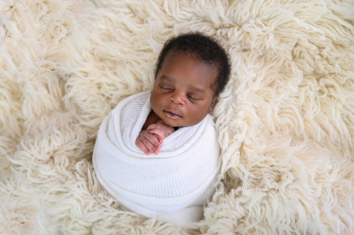 Black newborn boy wrapped in white on a cream background for his newborn photography session in Burlington, Ontario