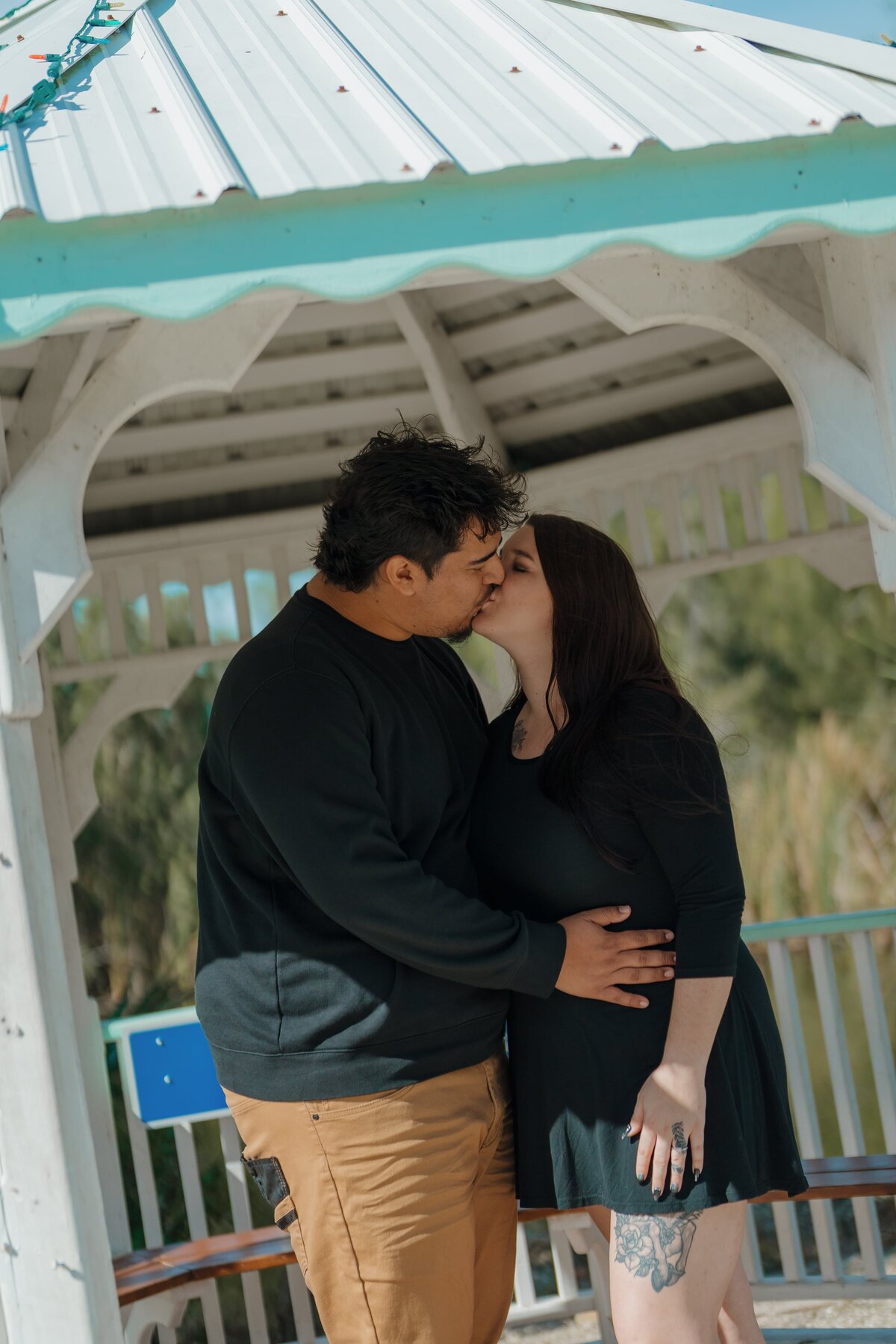 Couple sharing a kiss under a gazebo with a white roof, surrounded by greenery
