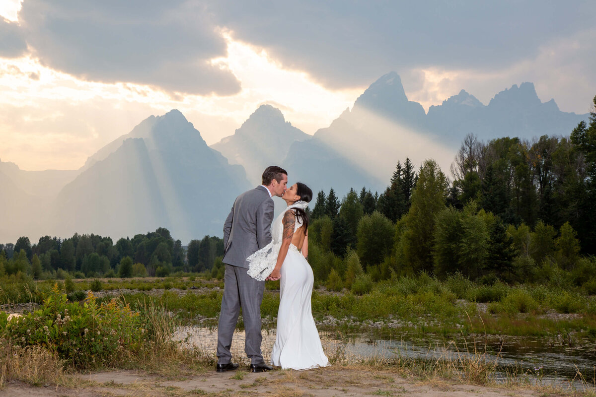 Bride and groom share a quiet moment at a Schwabacher’s Landing Elopement in Grand Teton National Park. Sunlight streams through dramatic clouds, highlighting the mountain peaks in this wedding portrait.