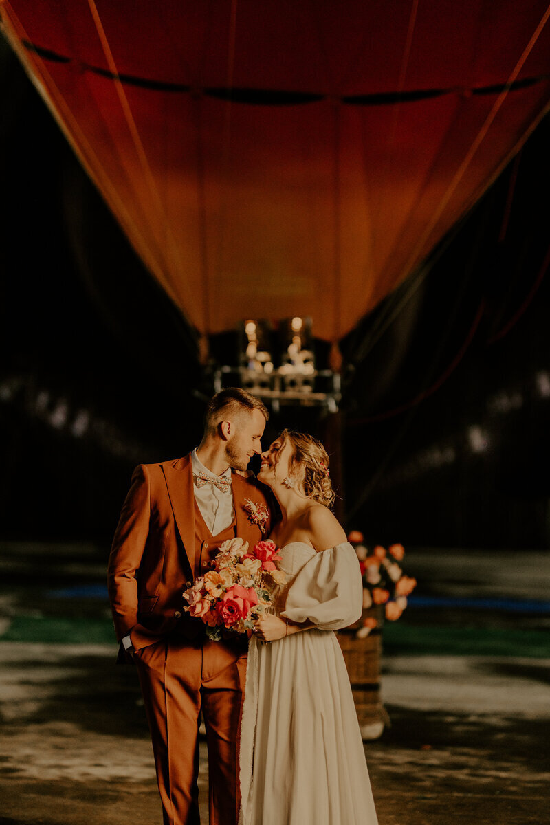 Couple de mariés, complices, les yeux dans les yeux. Marié en costume orange. Montgolfière en osier et ballon rouge en arrière plan pour une séance photo mariage.