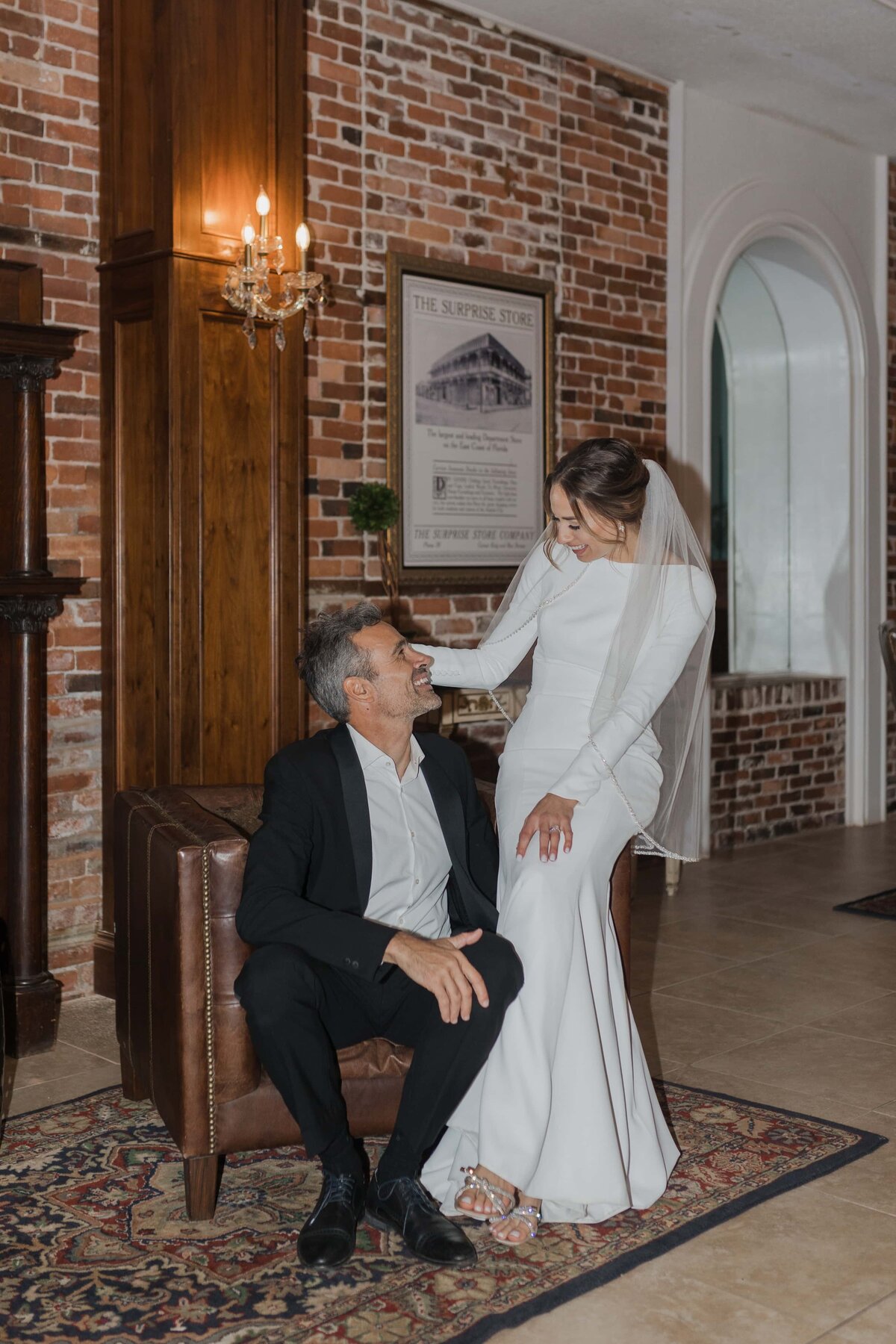 A candid moment between a bride and groom inside The White Room, a historic wedding venue in St. Augustine, Florida. The bride, dressed in a sleek long-sleeve gown with a chapel veil, gently rests her hand on the groom’s shoulder as they exchange a joyful look. The exposed brick wall, elegant chandelier, and warm tones of the room add to the romantic atmosphere. Captured by Phavy Photography in a documentary, true-to-color style that highlights the authentic connection and timeless setting.