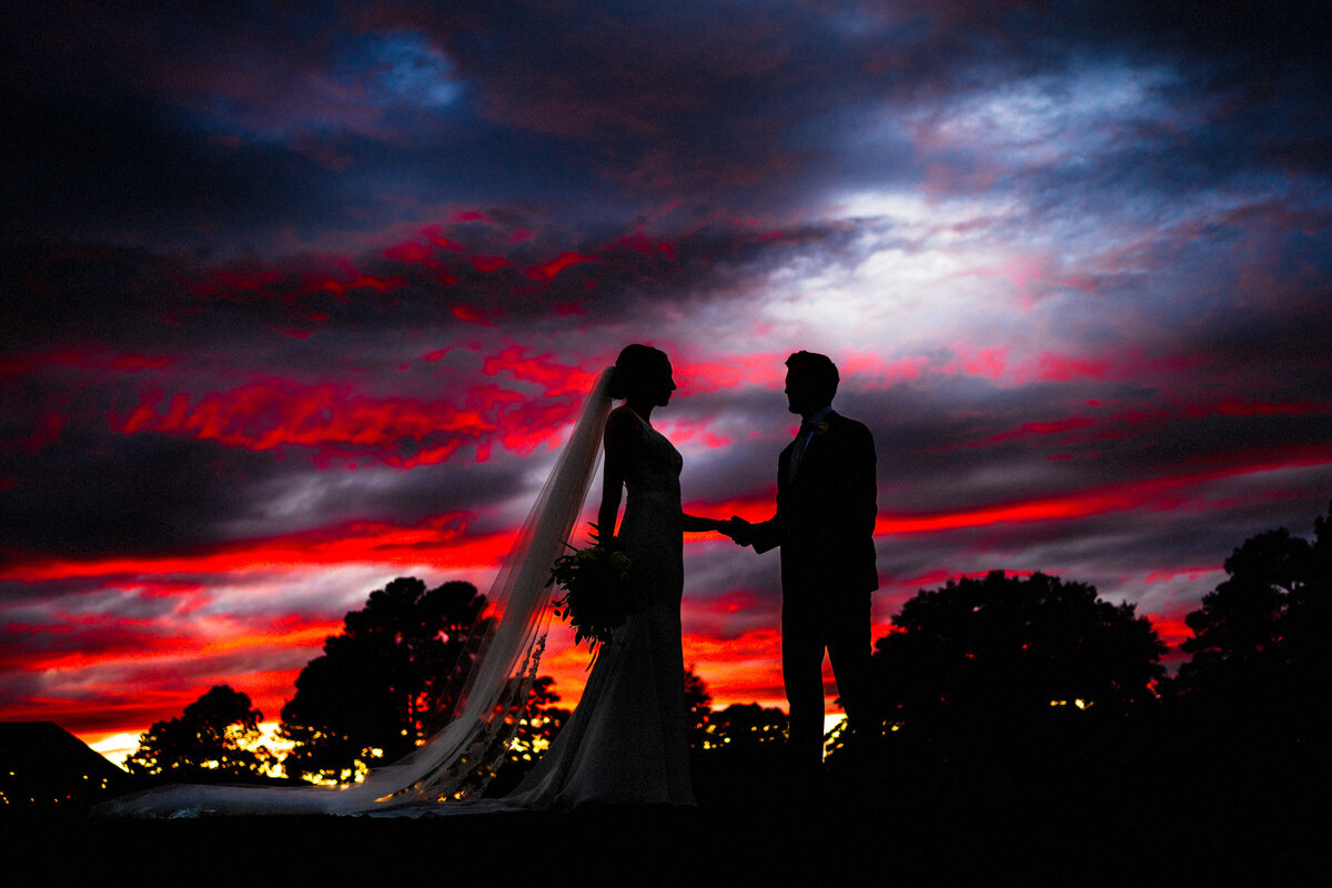 A bride and groom stand against a dramatic sunset sky, painted with vivid hues of red and blue, in a striking silhouette.