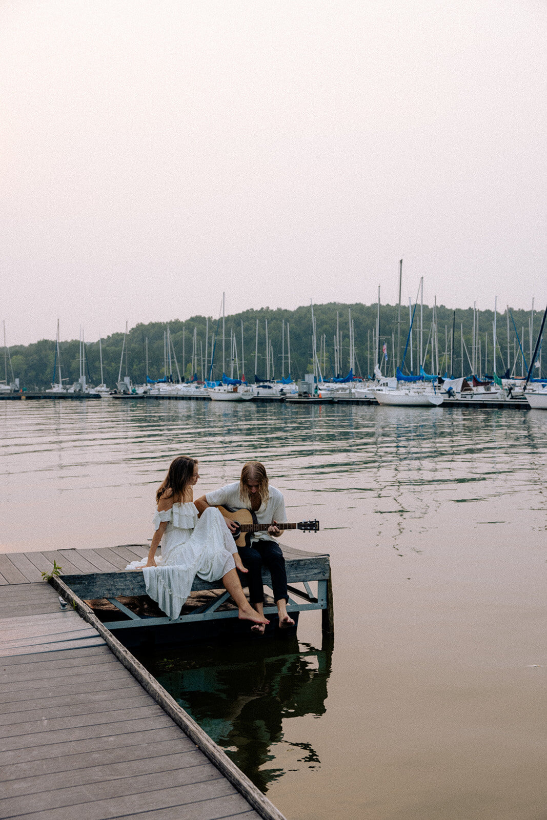 Engaged couple sits with guitar on a dock surrounded by sailboats at Hamilton Creek Marina in Nashville, TN.