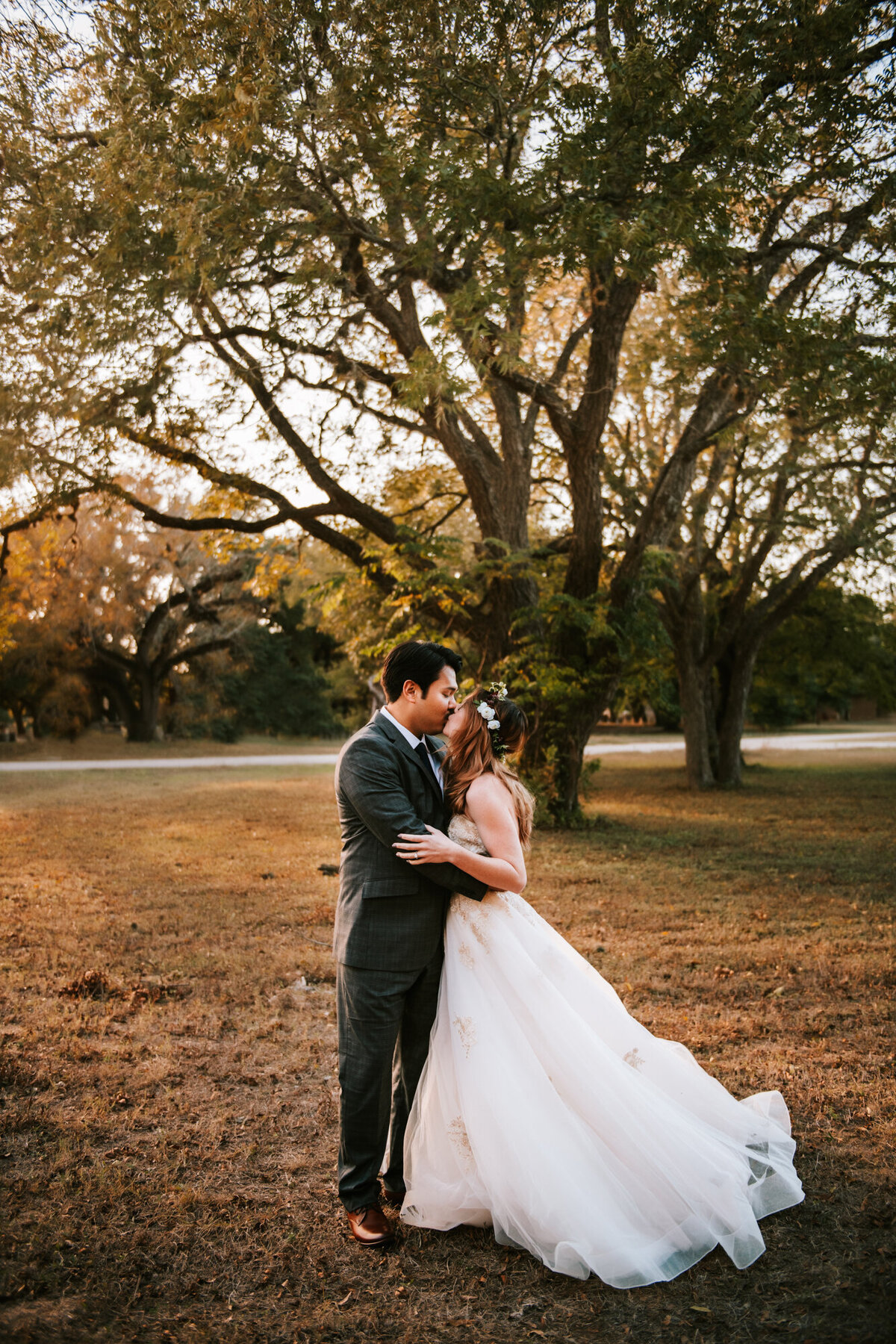 Couples Photography, Man in a blue blazer holding woman in a wedding dress and kissing her in front of large trees.