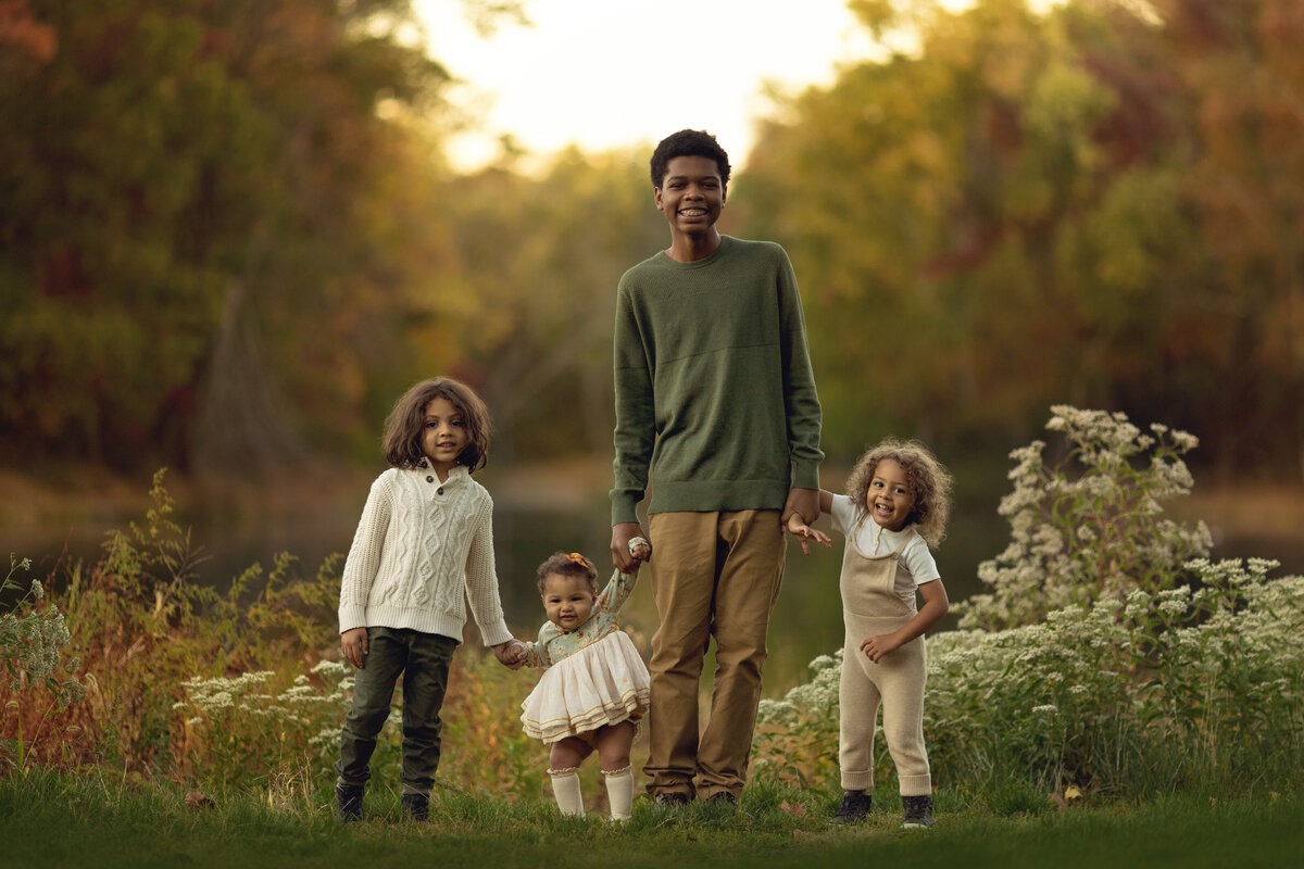 A teen boy in a green shirt holds hands and walks his three toddler siblings through a park at sunset posed by a New Jersey Family Photographer
