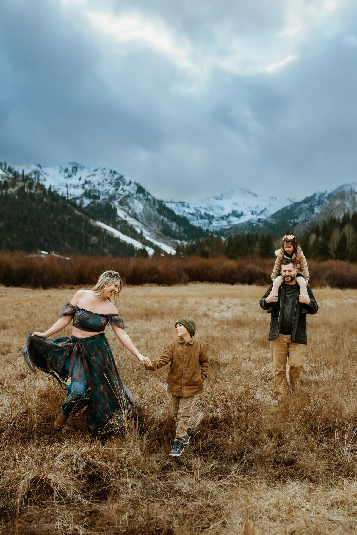 Mom is holding her son's hands looking at him and laughing as the dad walks behind them with their daughter on his shoulders in a field with mountains