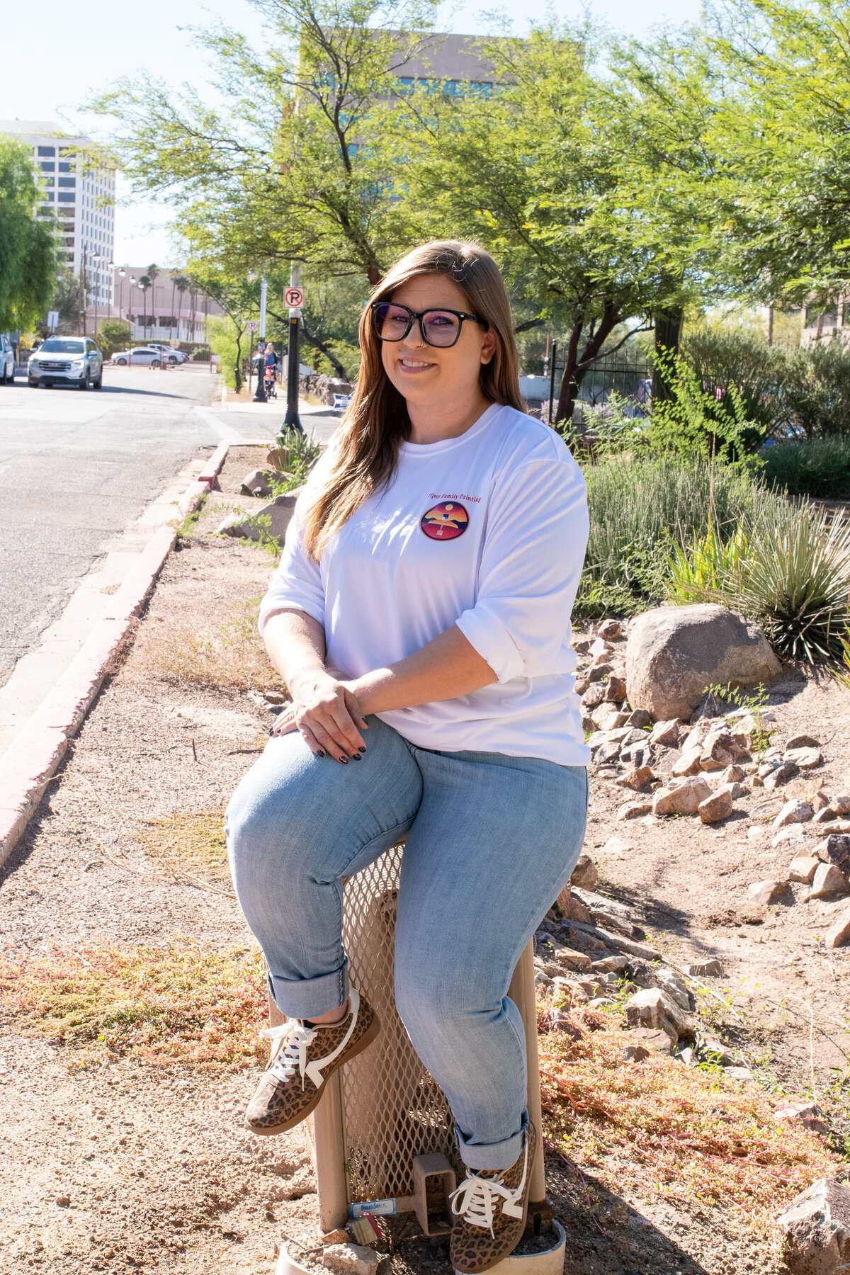 Woman sitting outdoors on a small metal seat along a city street, smiling toward the camera while wearing glasses and casual clothing, photographed by Vyrl Photo, showcasing Tucson brand photography.