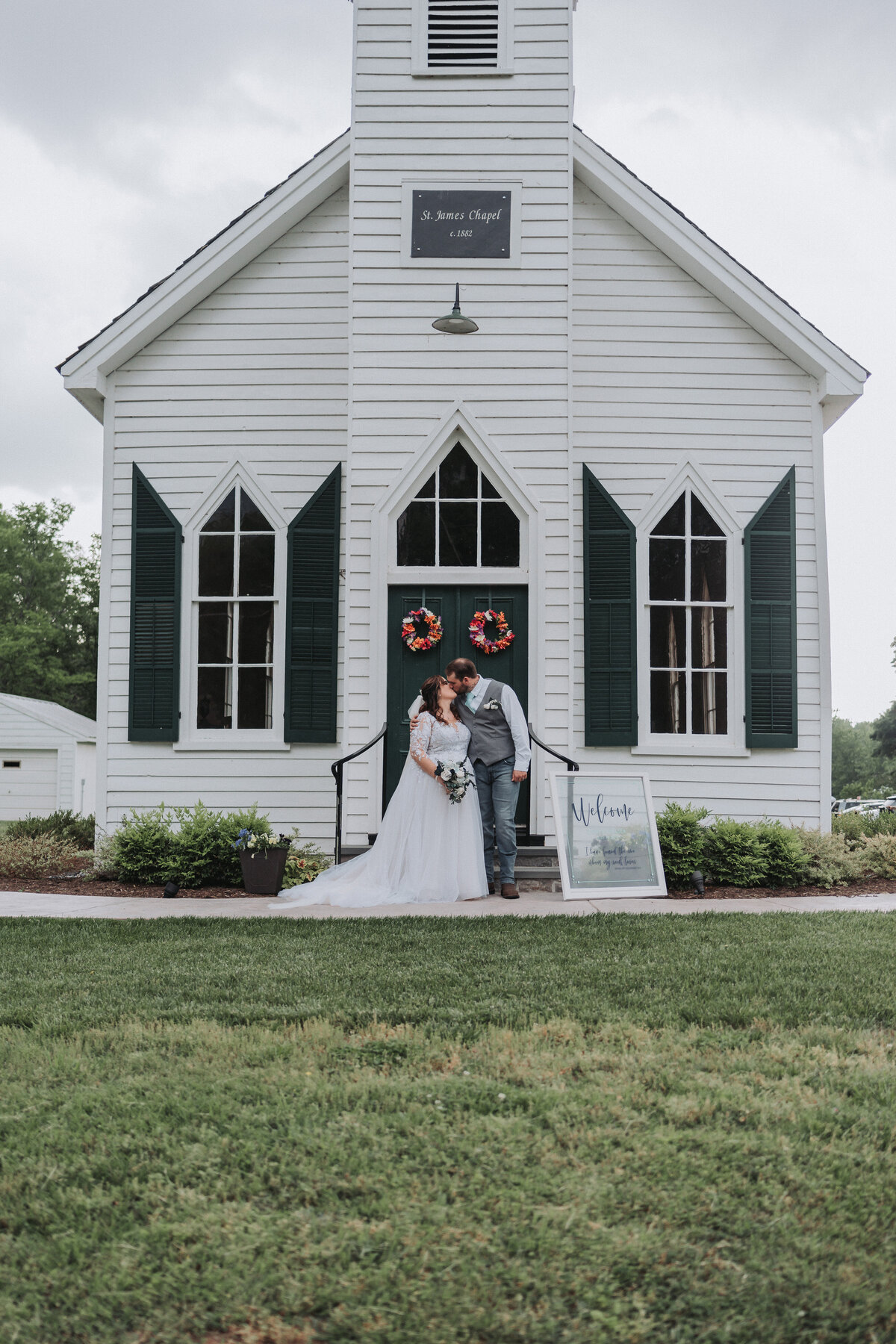 A happy wedding couple stands in front of the charming white St. James Chapel with dark green shutters at the Michaux-St. James Foundation in Virginia, beautifully captured by Quinn Photography LLC.