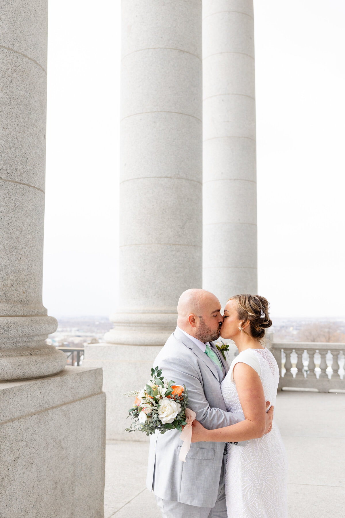 A bride and groom kiss while framed by towering marble columns, holding a soft floral bouquet.