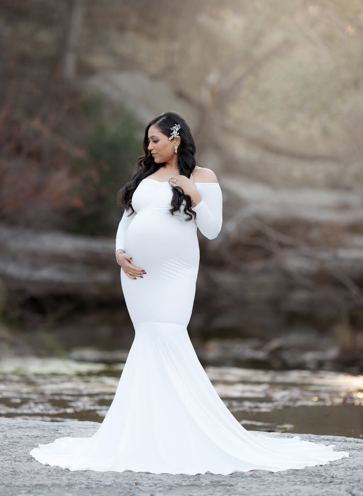 Expecting mother in a long white dress with jeweled hairpiece posing beside creek in Austin, TX