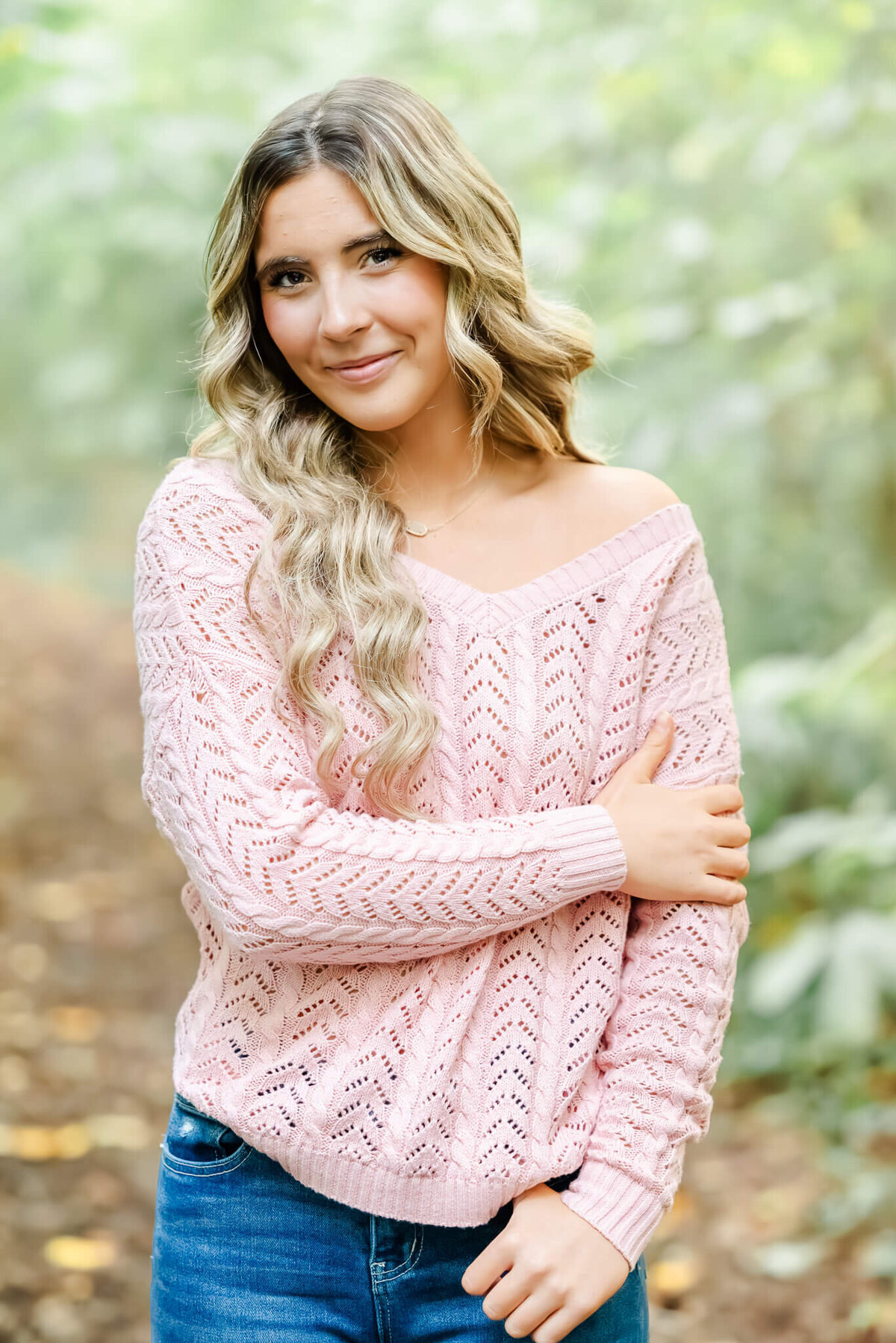 A high school senior stands on a path in the Chesapeake Arboretum during her senior session. She is wearing a pink sweater and jeans.