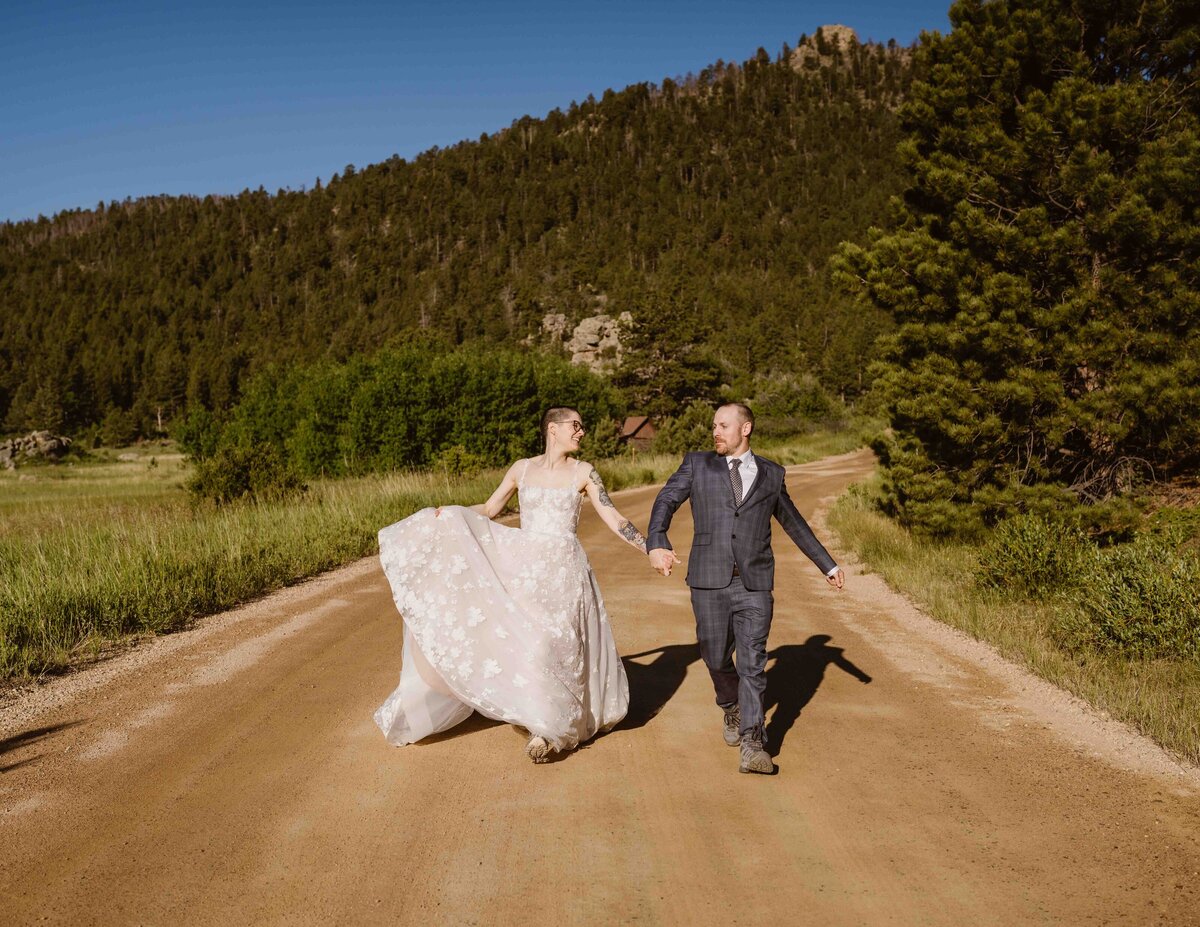  Bride and groom joyfully walk hand in hand down a dirt road in Estes Park, Colorado, surrounded by lush greenery and forested mountain hills under a clear blue sky.