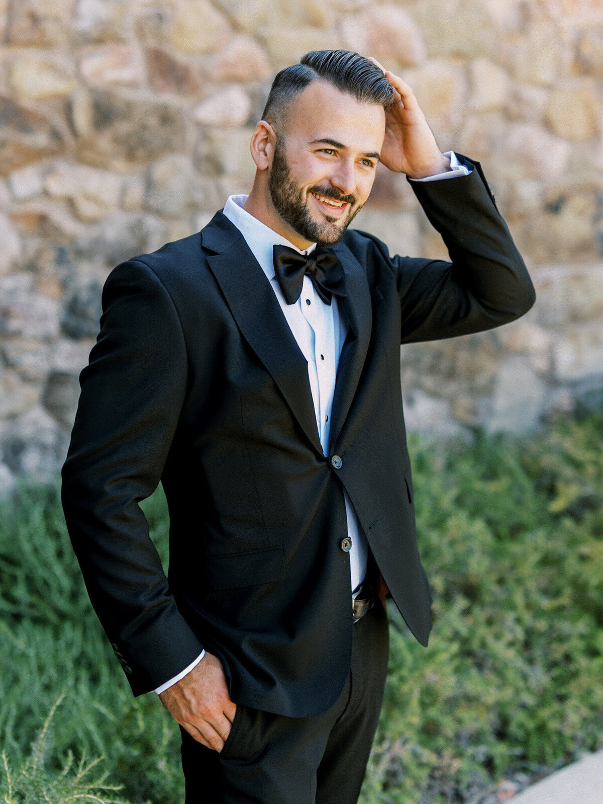 Groom in a classic black tuxedo, smiling confidently as he brushes his hair back, captured by Leana Myra Photography against a rustic stone wall and lush greenery in Santa Barbara.