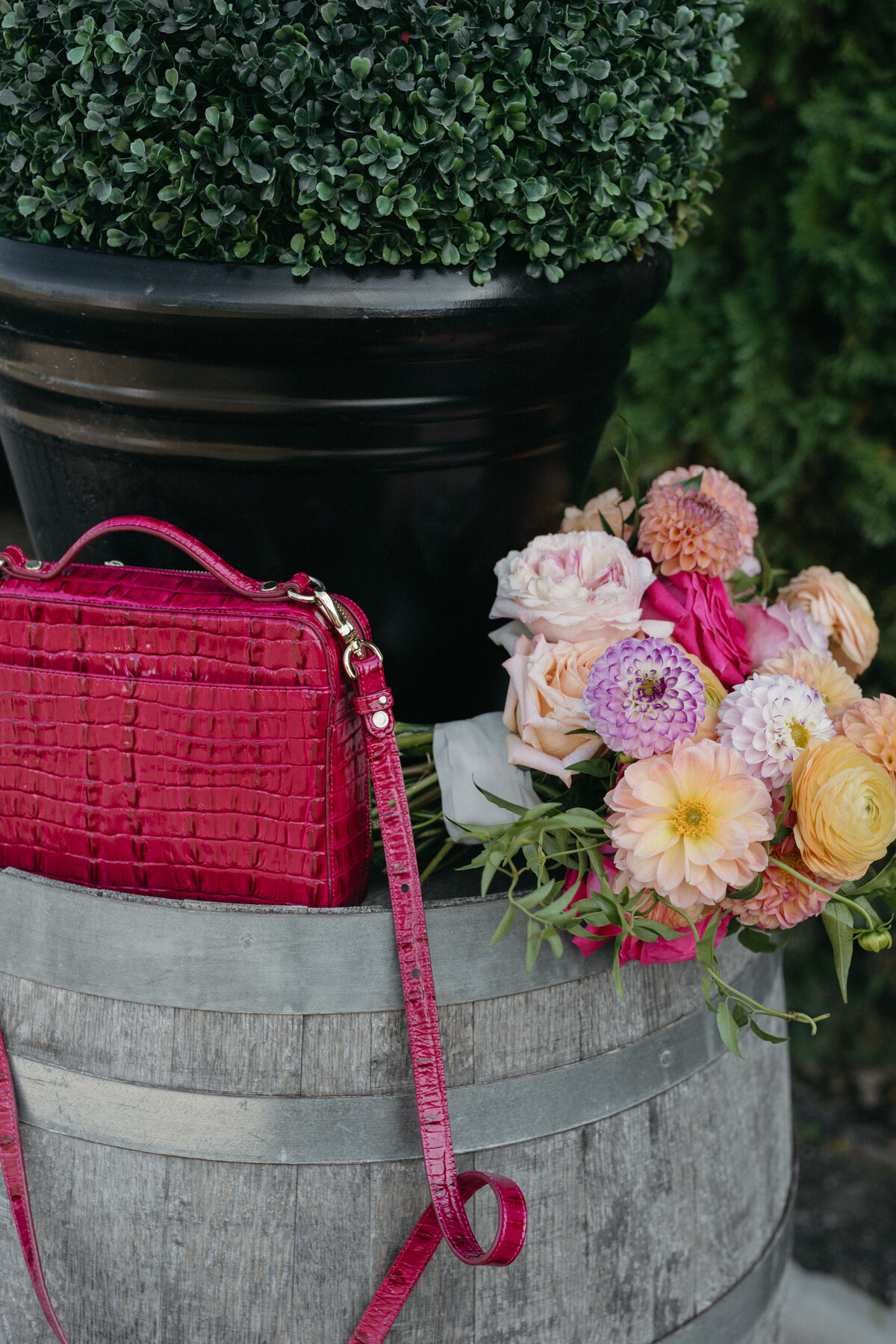 Bridal bouquet of dahlias, roses, and ranunculus in warm peach and pink hues resting on a rustic wine barrel beside a textured fuchsia purse.
