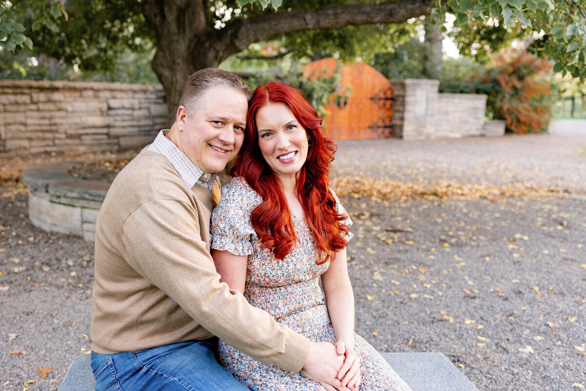 A smiling couple sits closely on a stone bench in a garden setting, surrounded by greenery and autumn leaves. The woman, with red hair, wears a floral dress, while the man wears a tan sweater and jeans.