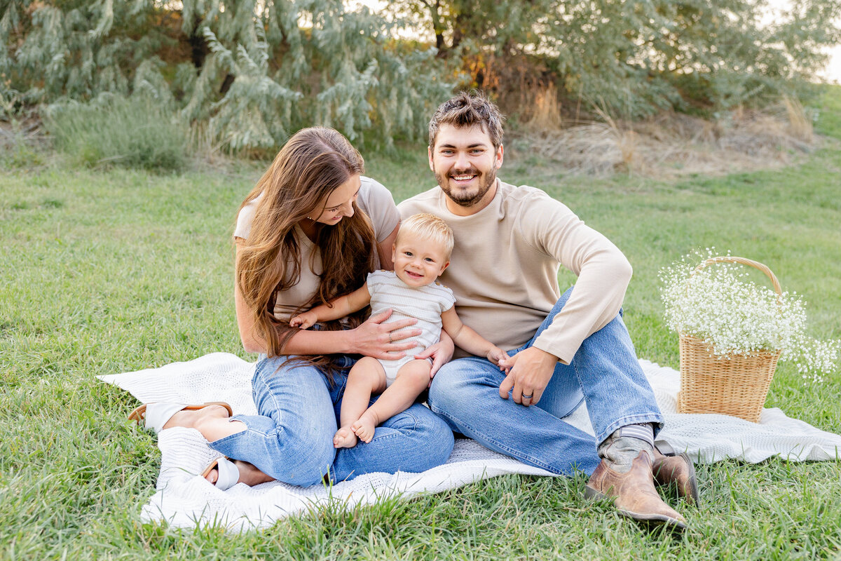 A family of three sits together on a blanket in a green meadow, with a wicker basket filled with flowers beside them. A beautiful family moment captured in Eagle Mountain, Utah.