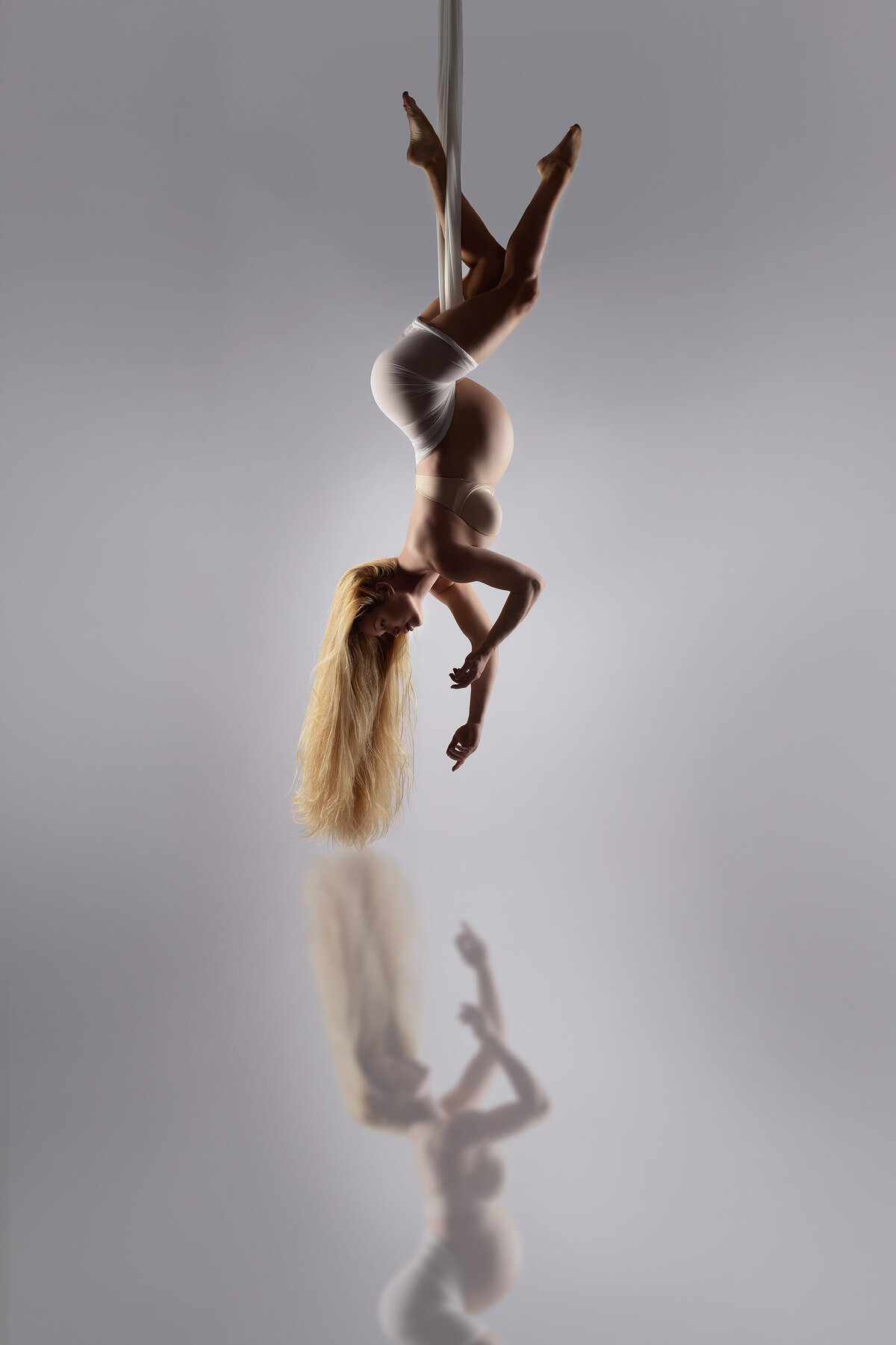 Woman gracefully suspended upside down from white aerial silks, her long blonde hair cascading toward the floor. Her serene expression and elegant pose are mirrored on the polished surface below.