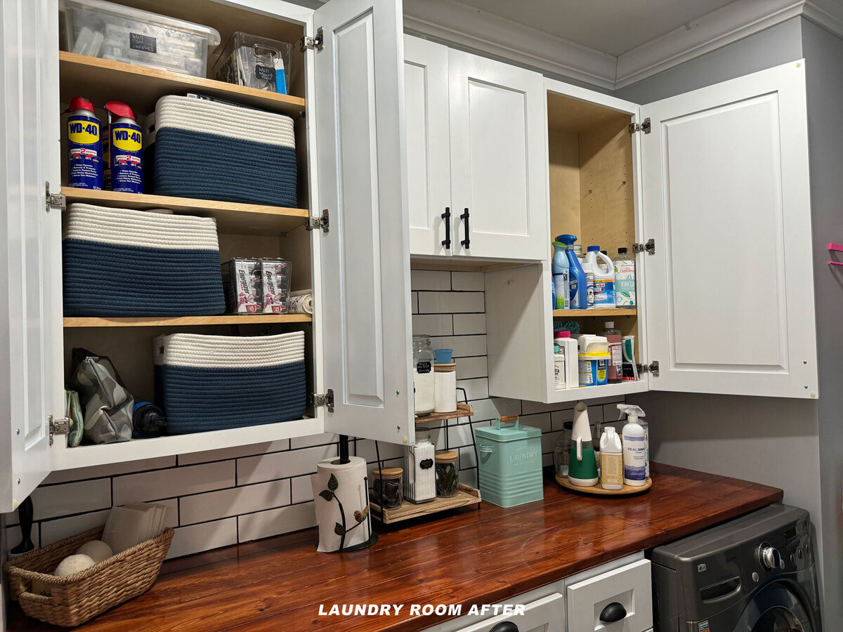 Organized laundry room with open white cabinets displaying baskets, cleaning supplies, and canned goods. A wooden countertop holds detergent, sprays, and other items. A washer is partially visible at the bottom right. Text reads "Laundry Room After.