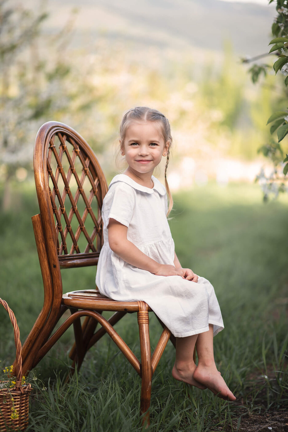 A toddler girl wears a white dress and sits on a wooden chair in the grass amongst and orchard. She is smiling. Portrait by Brittany Danielle.