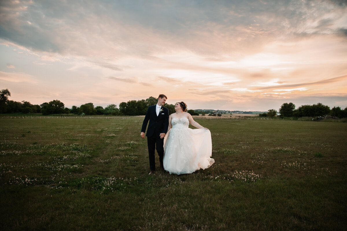 Newlyweds hold hands while walking through a pasture at sunset