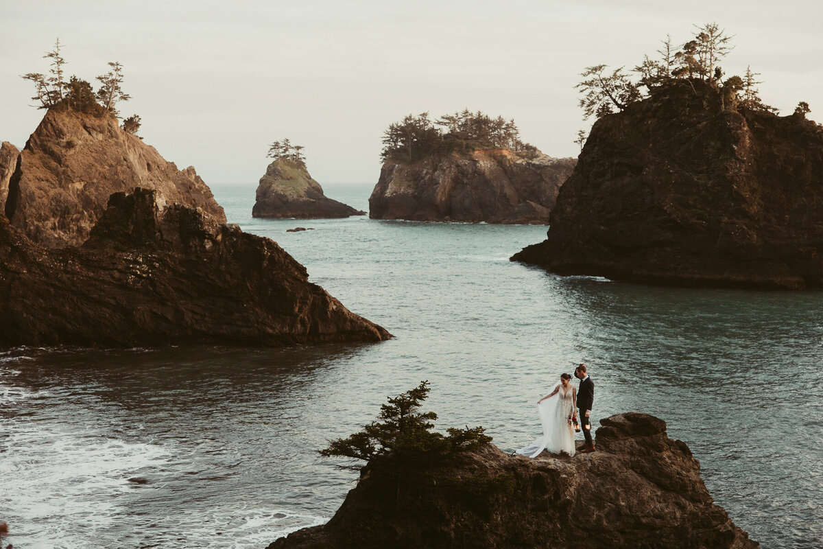 An epic wide shot of a bride in a dress and a groom in his soon standing on a cliffside overlooking the pacific ocean wiping tears while seeing his bride for the first look from a Vancouver WA Wedding Photographer