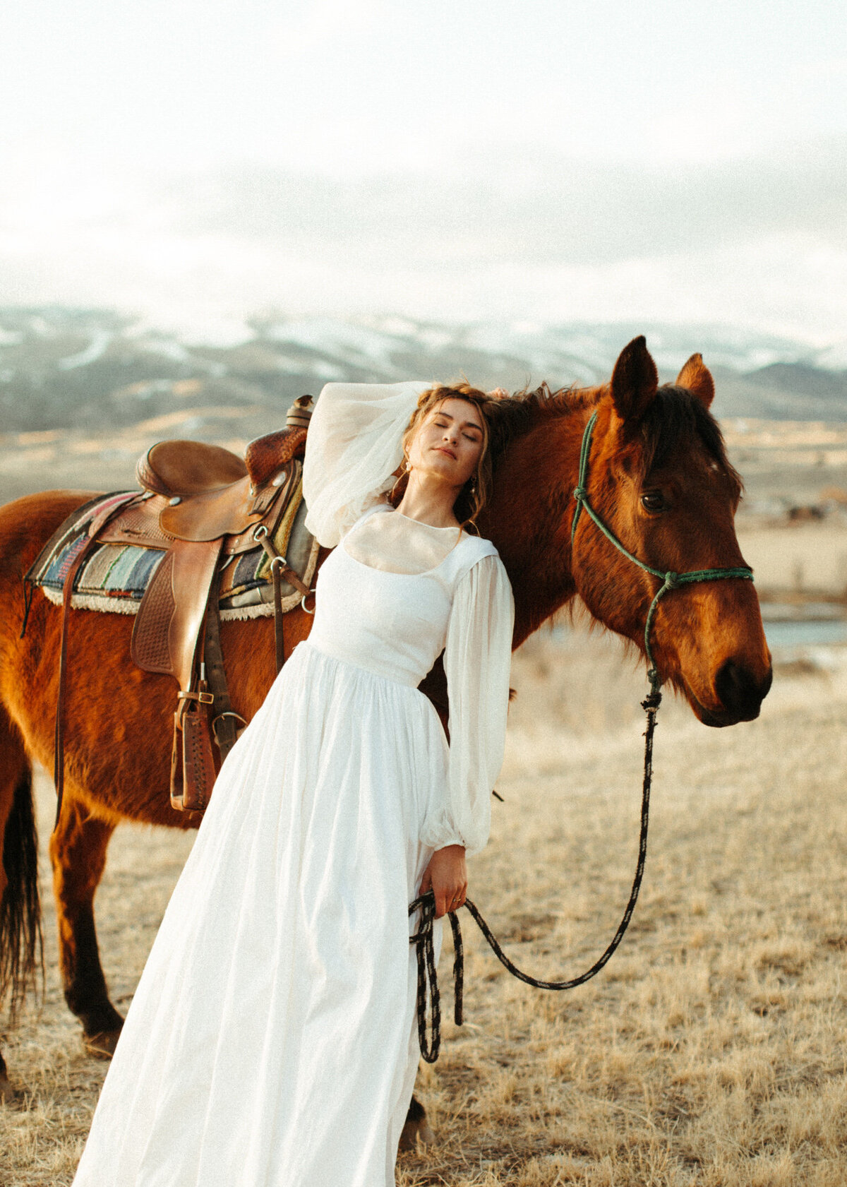 Bride in wedding dress leaning back against a horse closing her eyes with snowy mountains behind them