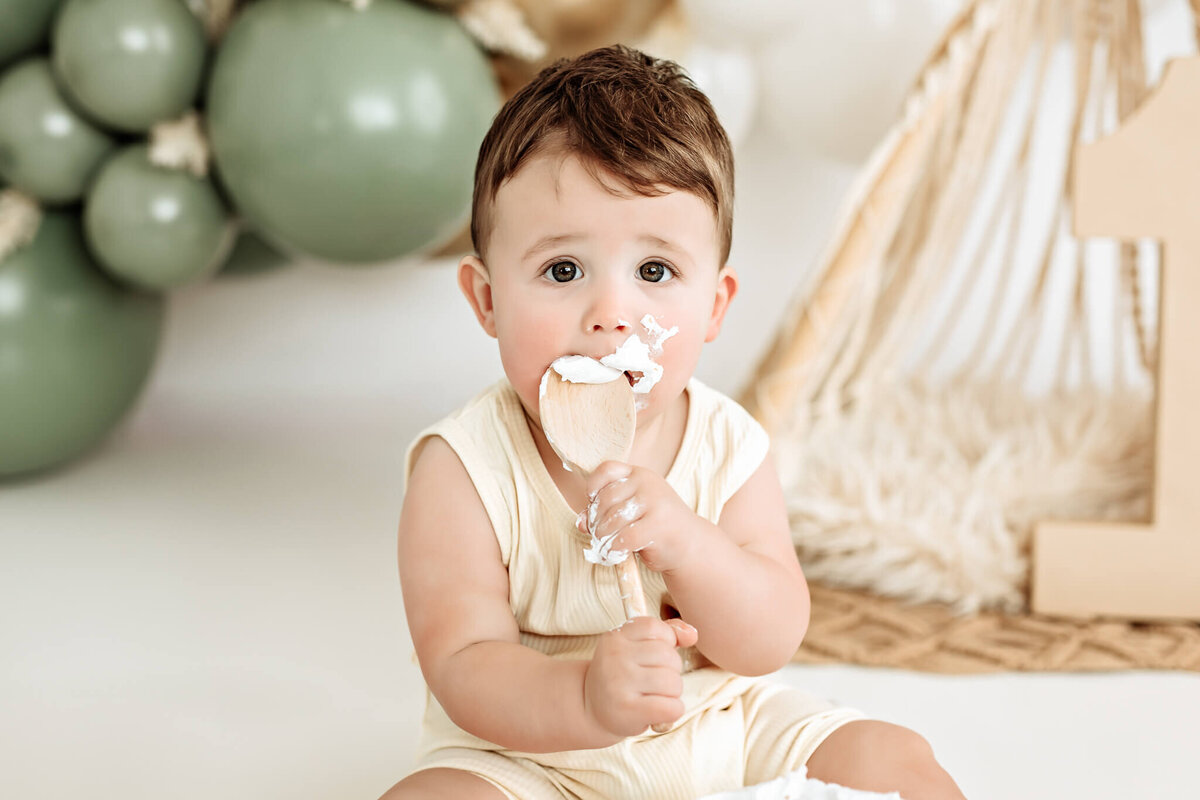 A baby sitting on a soft surface holds and bites a cake-covered spatula. Green balloons and a large wooden number one are in the background, suggesting a first birthday celebration.