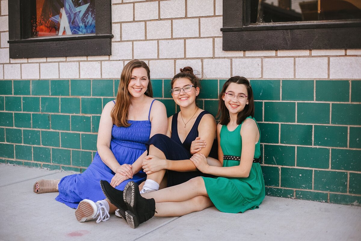 mother-and-two-daughters-in-colorful-dresses-sitting-against-green-brick-wall-smiling-during-urban-broadripple-indianapolis-family-session