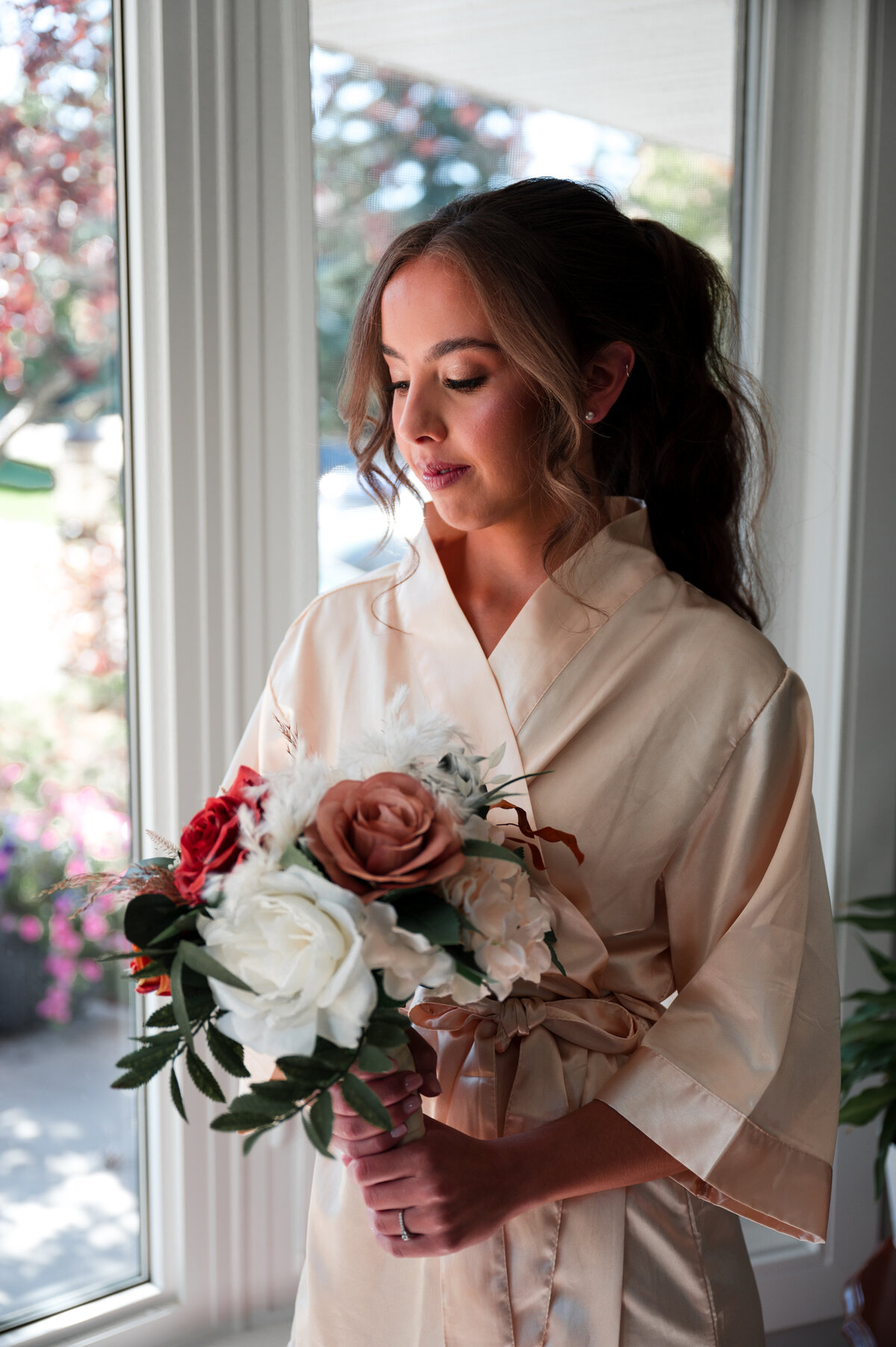 bride holding a boquet of flowers standing by windows in a white robe, getting ready for her wedding