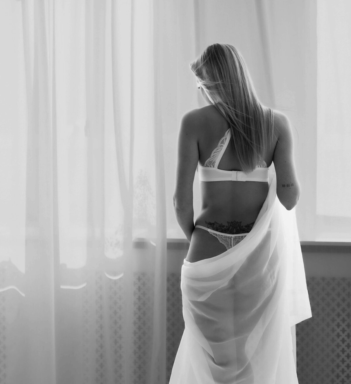 Staande boudoirpose bij het raam. Model draagt witte lingerie icm een wit laken.