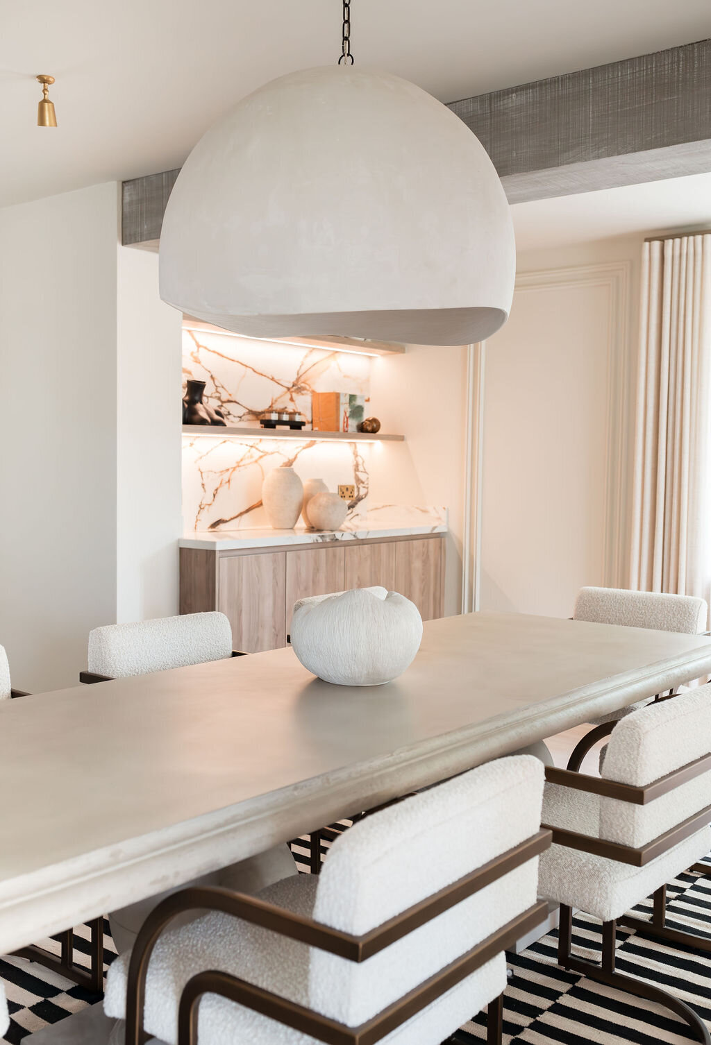 A modern dining room with a large rectangular wooden table surrounded by cushioned chairs. Behind the table is a backlist marble bar, emitting a soft glow. A large dome-shaped pendant light hangs above the table, and a black and white striped rug lies underneath.