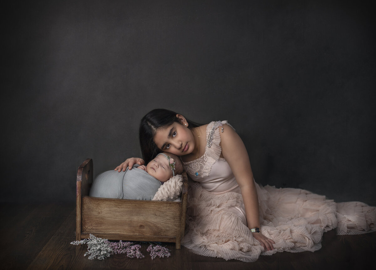 New born girl is swaddled and laying in a bed while older sister  gently rests beside in a light pink dress. Photo taken in Sonia Gourlie Fine Art Photography 's Studio.