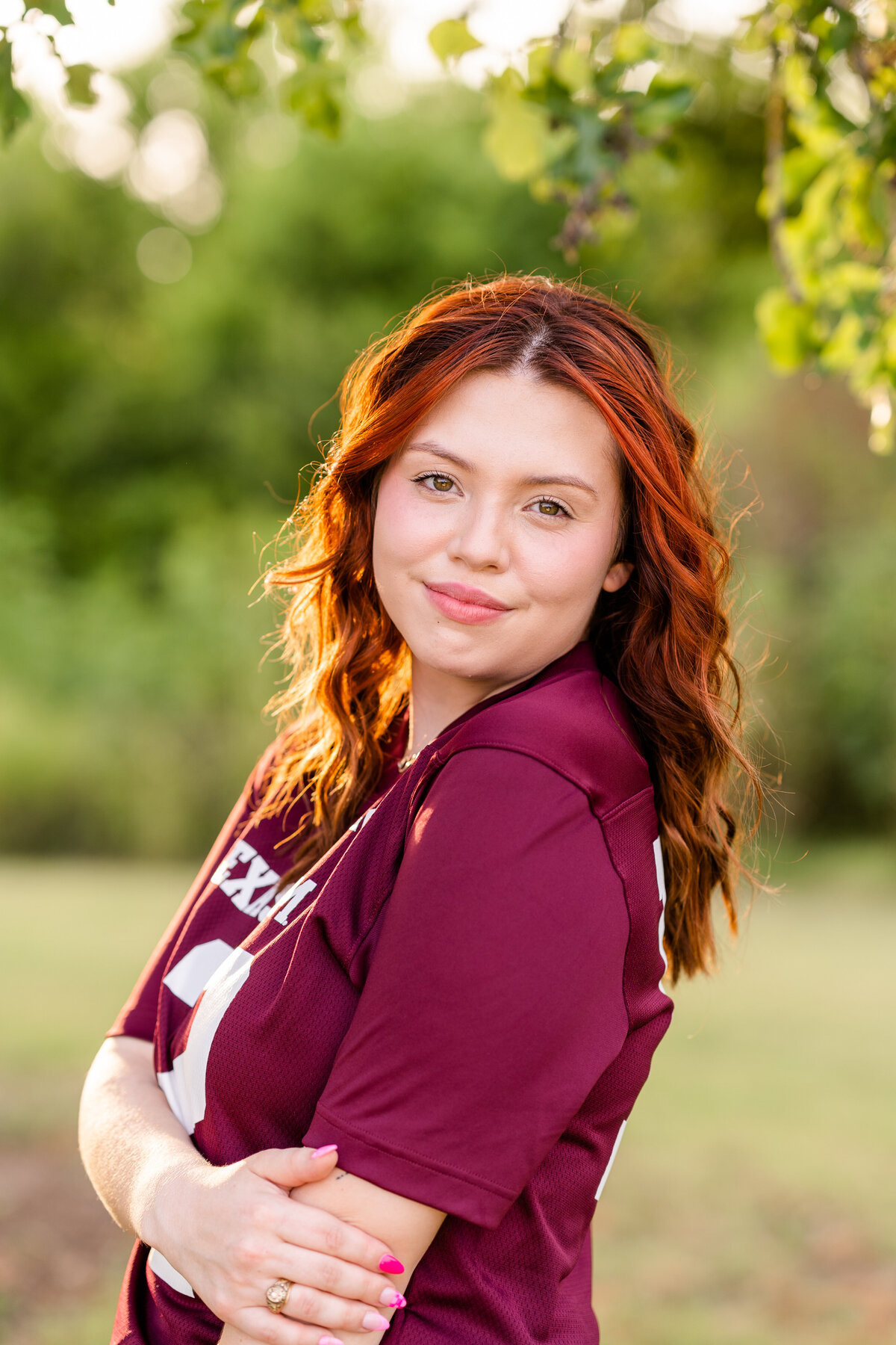 Texas A&M senior girl looking over shoulder and smiling at camera wearing maroon jersey and holding elbow surrounded by nature at sunset at Leaching Teaching Gardens