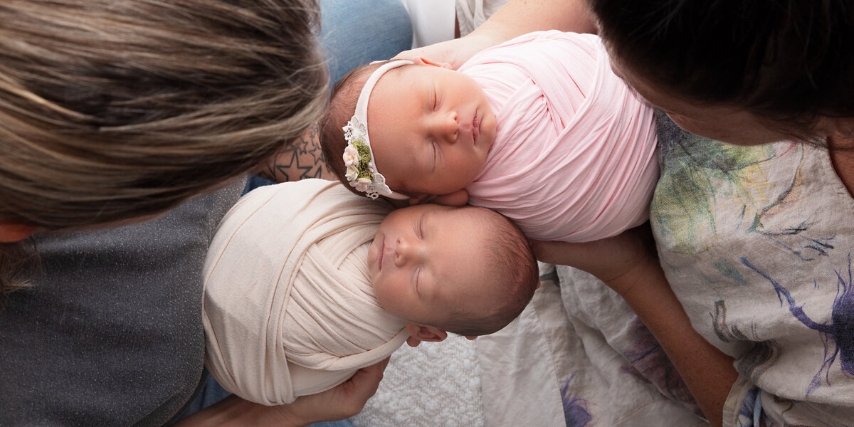 Newborn twins wrapped in cream and pink swaddles, cradled in their parents’ arms as the parents look lovingly down at them.