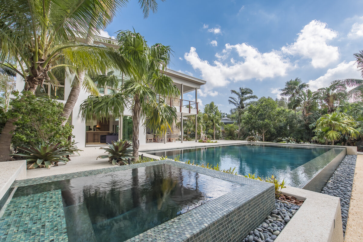 Feature photo for the Gordon Glory gallery. Backyard infinity pool and hot tub with blue teal tiles, large palm trees, and the large modern all white house.