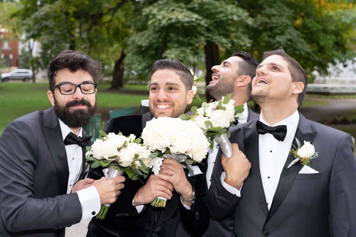 Groomsmen humorously pose while holding bouquets, adding a playful and light-hearted touch to the wedding portraits. This image captures their fun personalities and camaraderie, contrasting with the traditional aspects of the wedding and showcasing a unique and memorable moment.