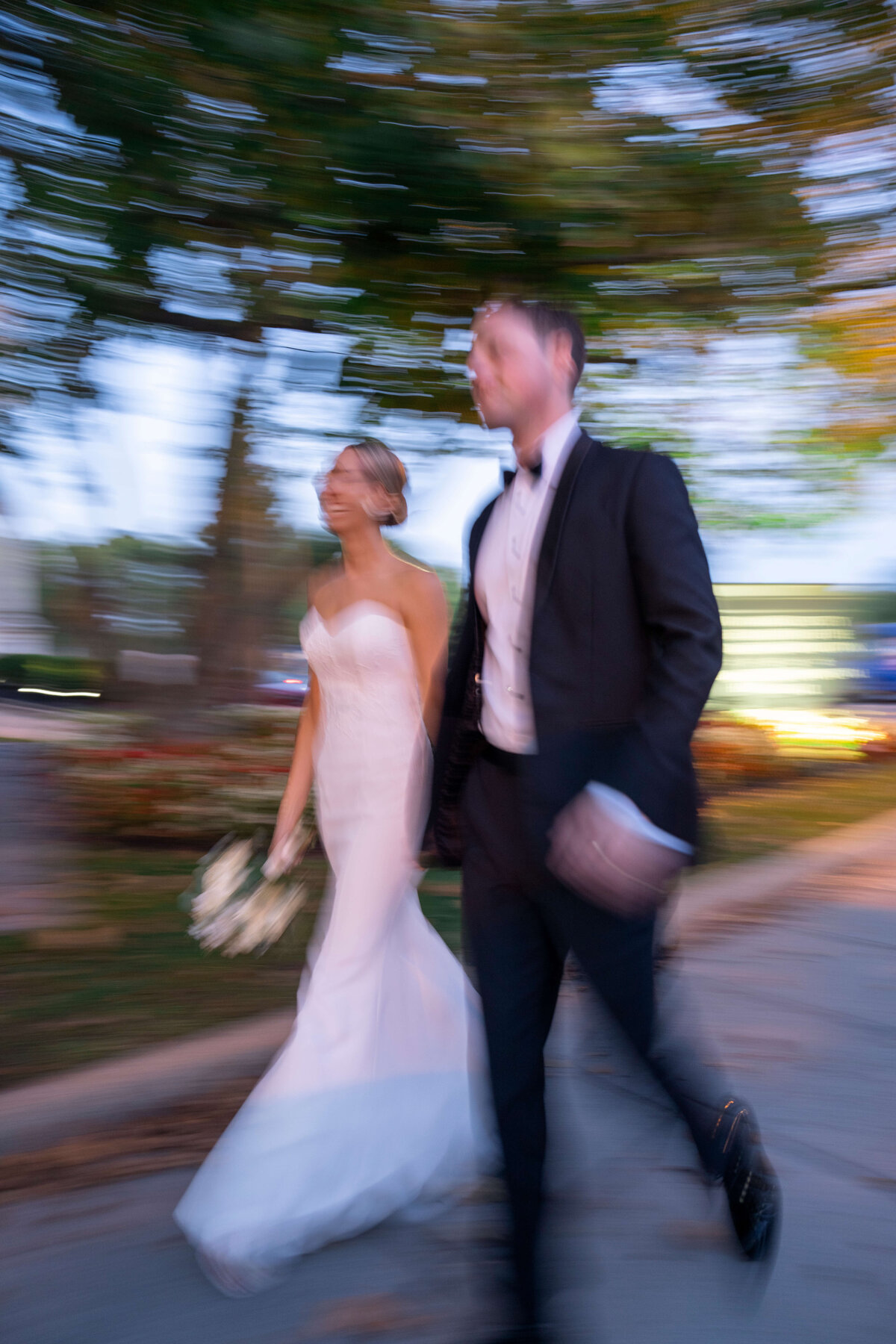 A bride in a white dress and a groom in a black suit walk together. The background is blurred, suggesting motion, with trees and blurred lights in the distance. The bride holds a bouquet of flowers.