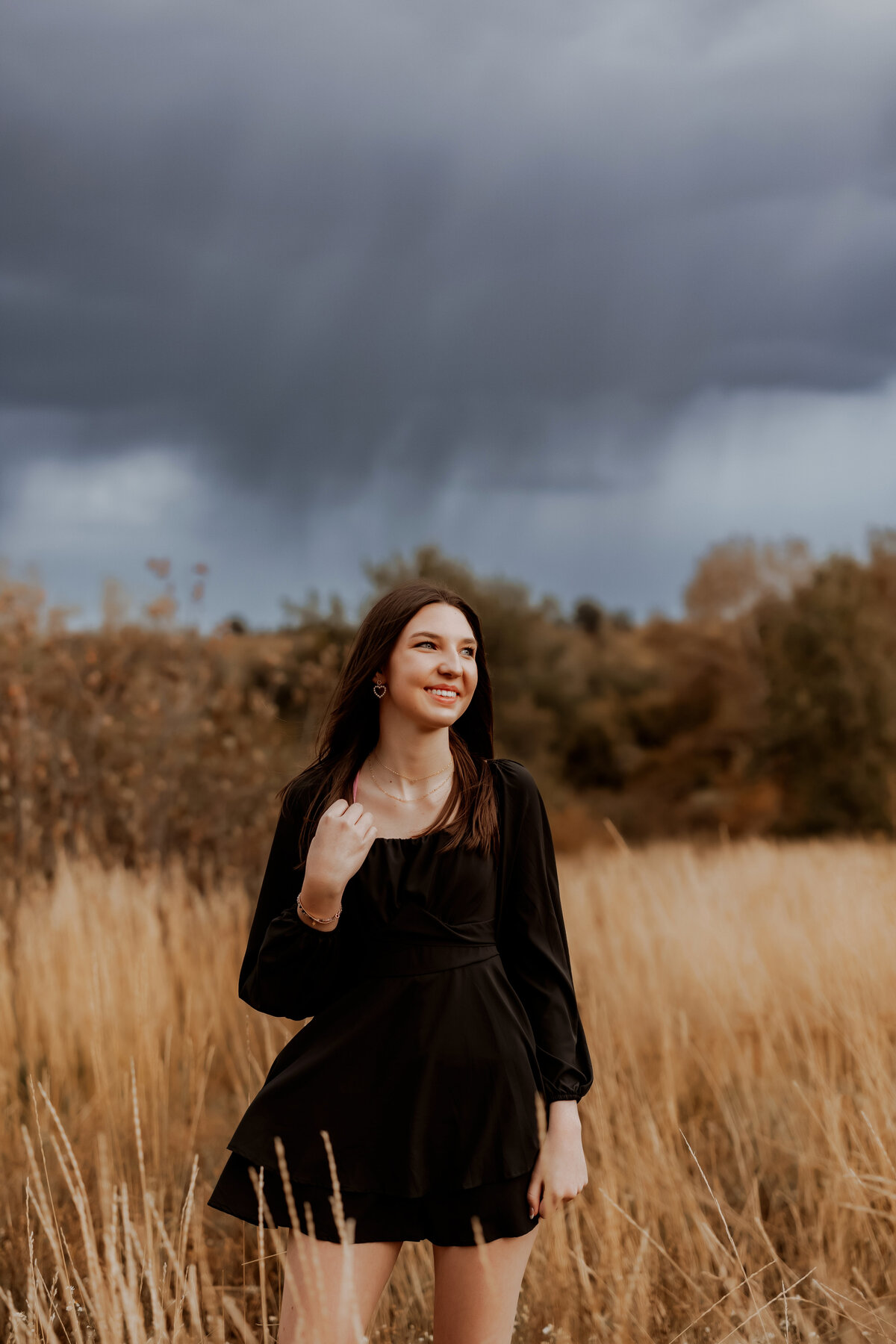 high school senior girl in black dress stands in wheat field with dark clouds in the back ground