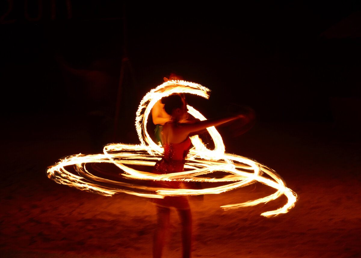 A woman in a red dress dances with fire on a beach at night providing party entertainment.