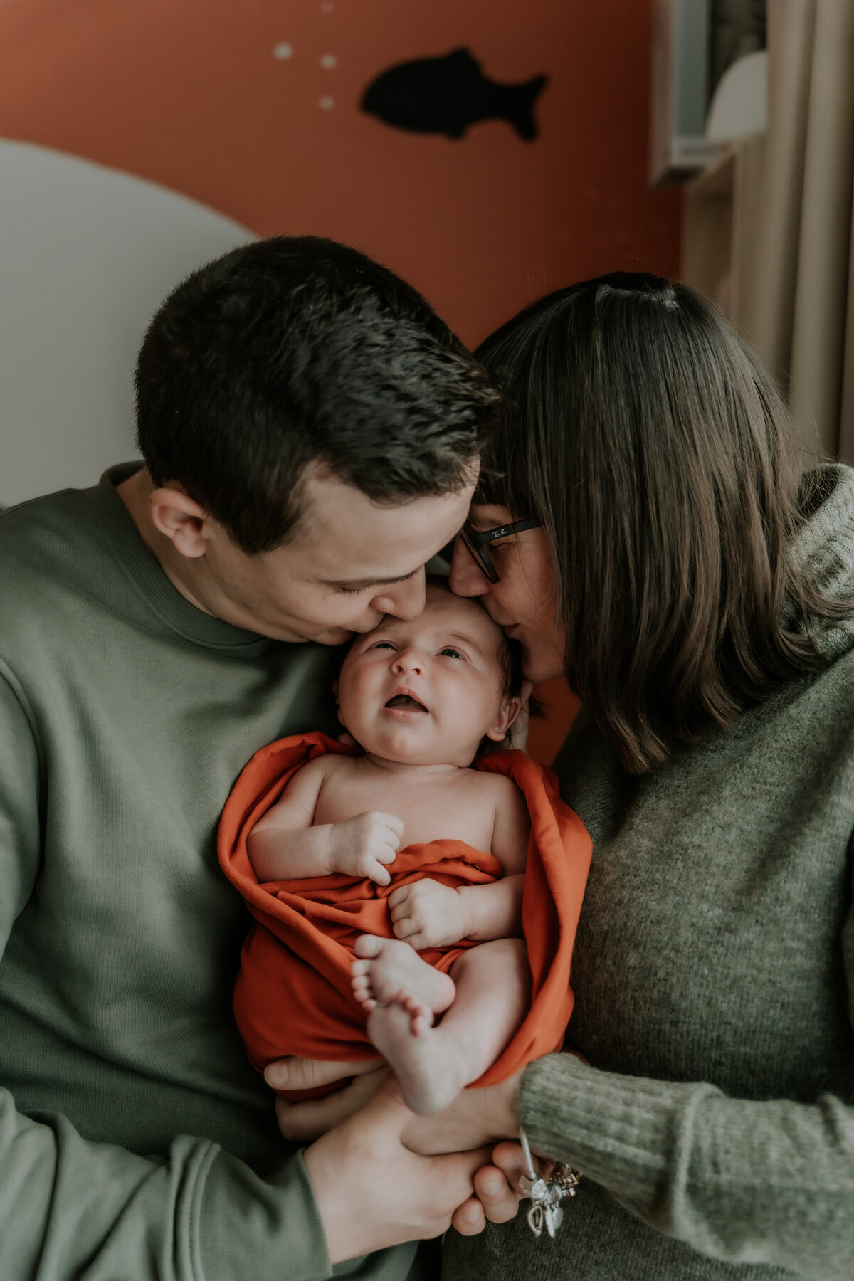 Newborn genietend in de armen van de ouders, ouders geven beiden een kusje op het hoofdje. De newbornshoot ging door in Deurne Antwerpen.