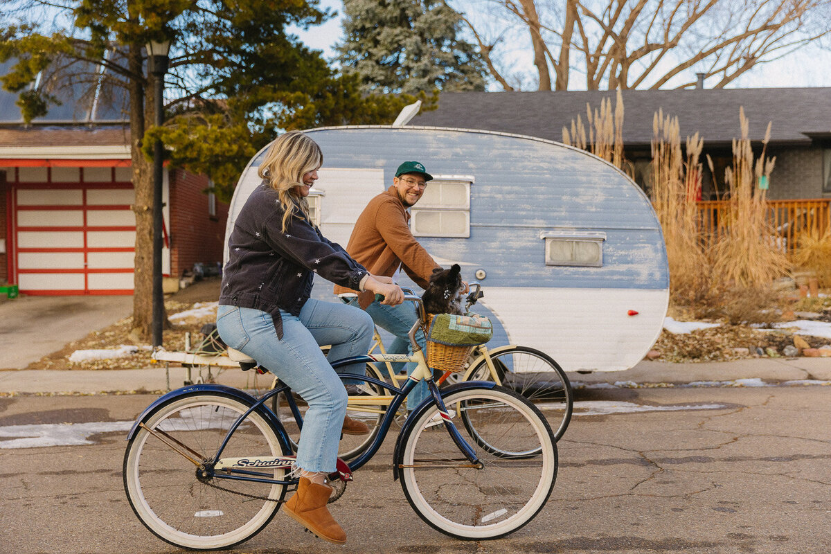 Couple riding bikes for their engagement photos in their neighborhood in front of the camper van they renovated together.
