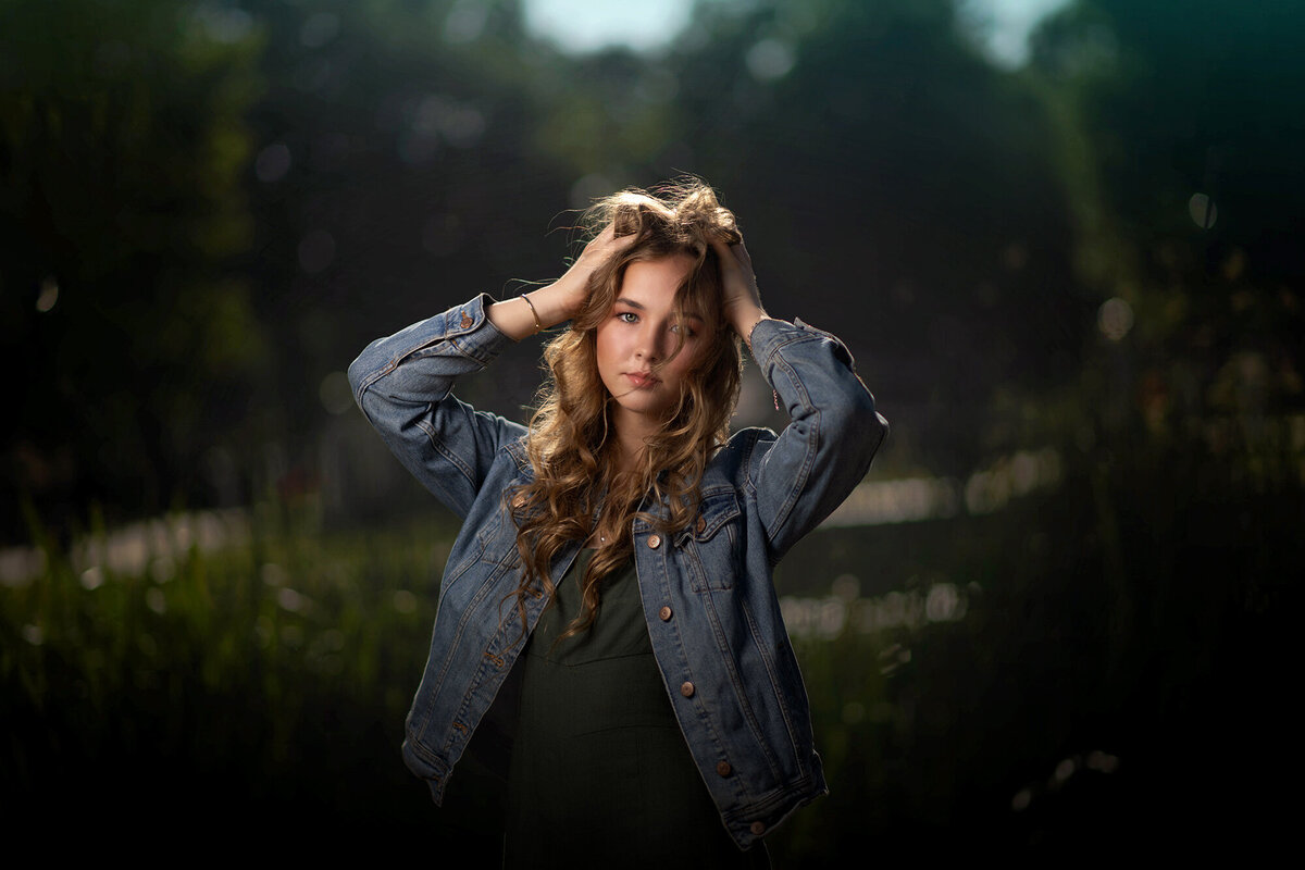 A high school senior bunches her hair on her head while standing in a field in a denim jacket