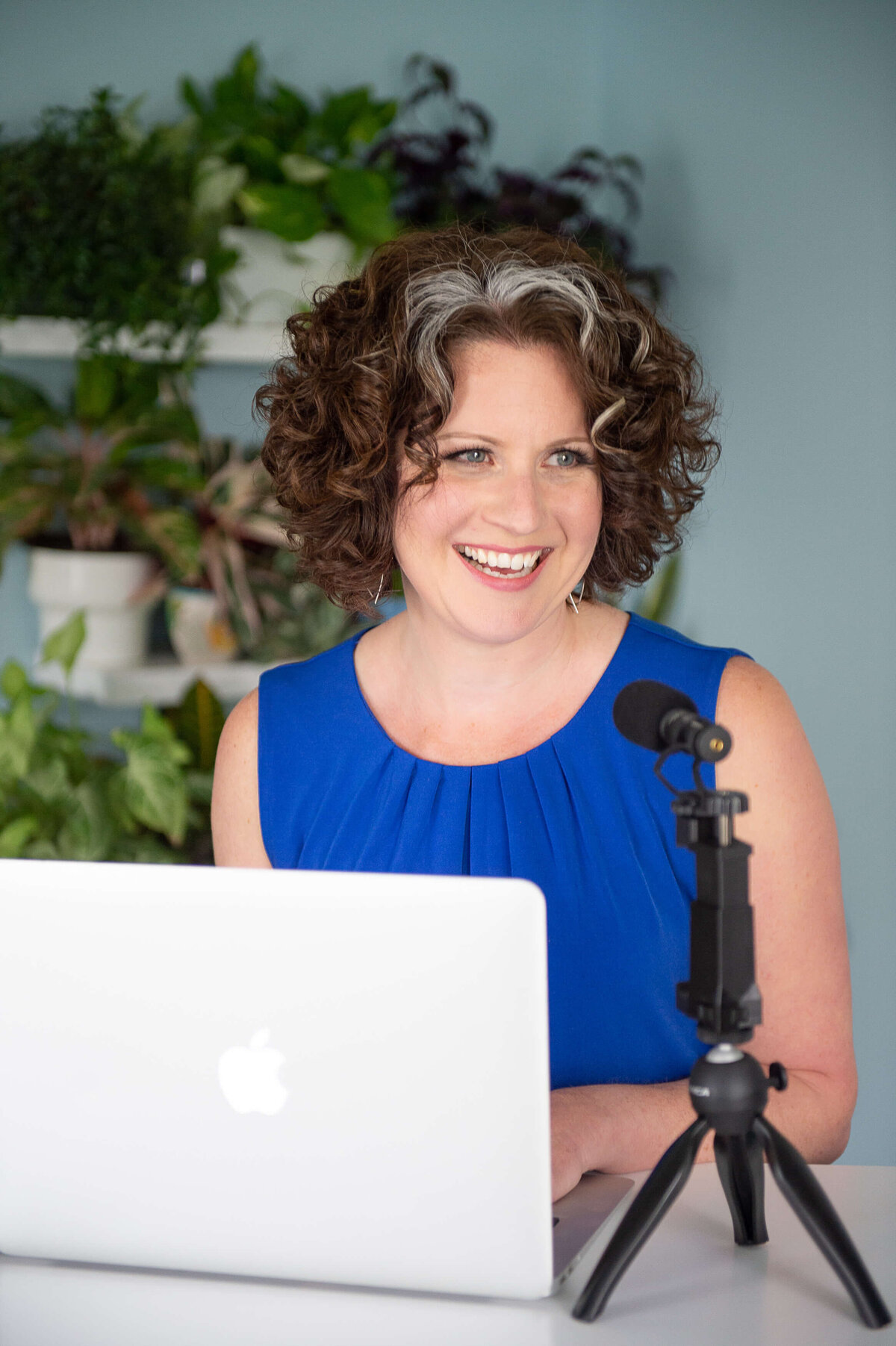 a woman in a blue blouse talking and laughing into her podcast microphone with her laptop in front of her.  Captured on-location by Ottawa Branding Photographer JEMMAN Photography COMMERCIAL