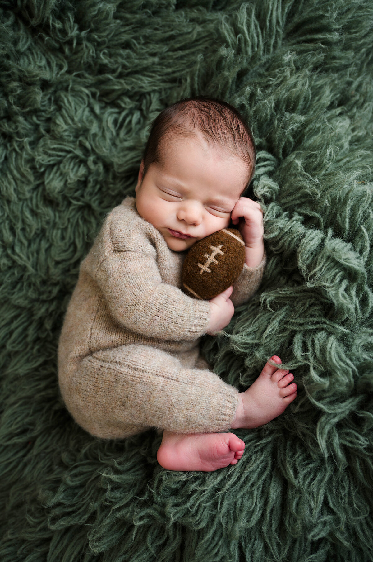 This heartwarming newborn baby boy photography session in Pittsburgh captures the serenity of a sleeping baby holding a small football. Wrapped in a cozy knitted onesie and resting on a soft green fur backdrop, this photo celebrates the precious first days of life.