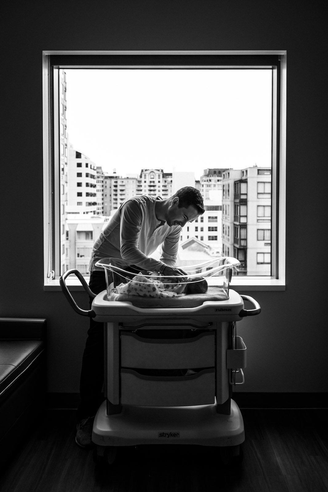 dad standing by window and leaning over newborn in hospital bassinet