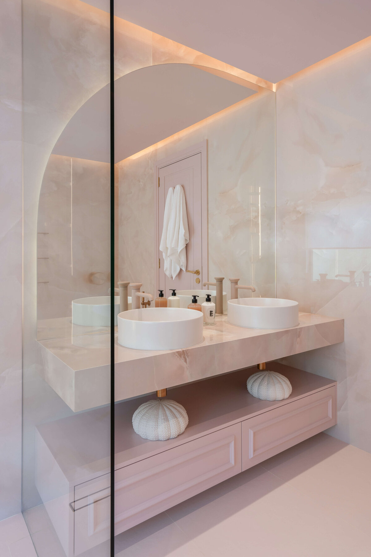 Double sinks in a light pink marble bathroom. The cabinets are pink, the counters marble pink, with matte pink spouts and white bowls. The mirror above the two sinks has one rounded  corner at the top left.